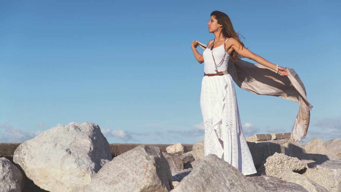 femme sur des rochers portant un robe longue blanche et tenant un foulard